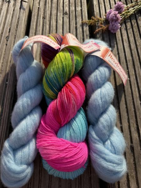 Handgefärbtes Garnset „Sommerregen“ von Herzfasern auf einer Holzbank im Sonnenlicht fotografiert. In der Mitte liegt ein bunter Strang Herzfasern No. 11 in der Farbe „Regenbogenkuss“ mit kräftigem Pink, Türkis, Grün und Violett. Daneben liegen zwei fluff
